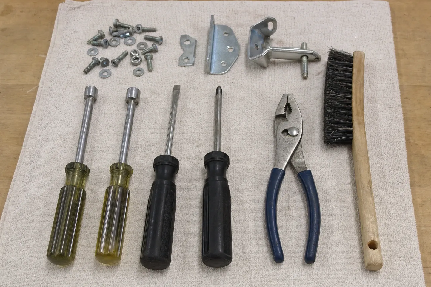 An L-bracket with a threaded stud, a four-hole mounting plate and a small tab laid above two nut drivers, a flat and a Phillips driver, pliers and a wooden-handled brush