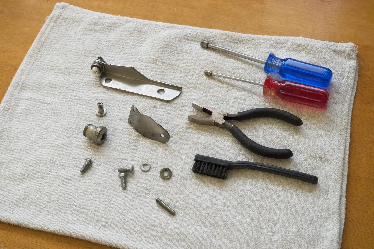 A bent lever arm with its pivot roller, a small bracket, a stepped bushing, a shoulder screw, two washers and a roll pin on a towel with needle-nose pliers and two nut drivers