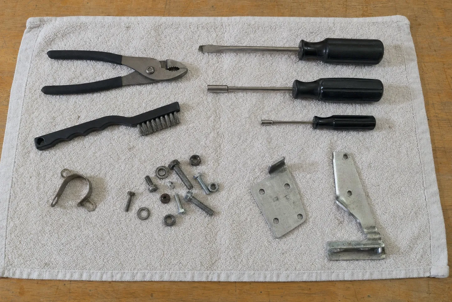 A curved strap clamp, a cluster of bolts, nuts and washers, a four-hole plate and an angled steel bracket set out with slip-joint pliers, a small brush and three black-handled drivers