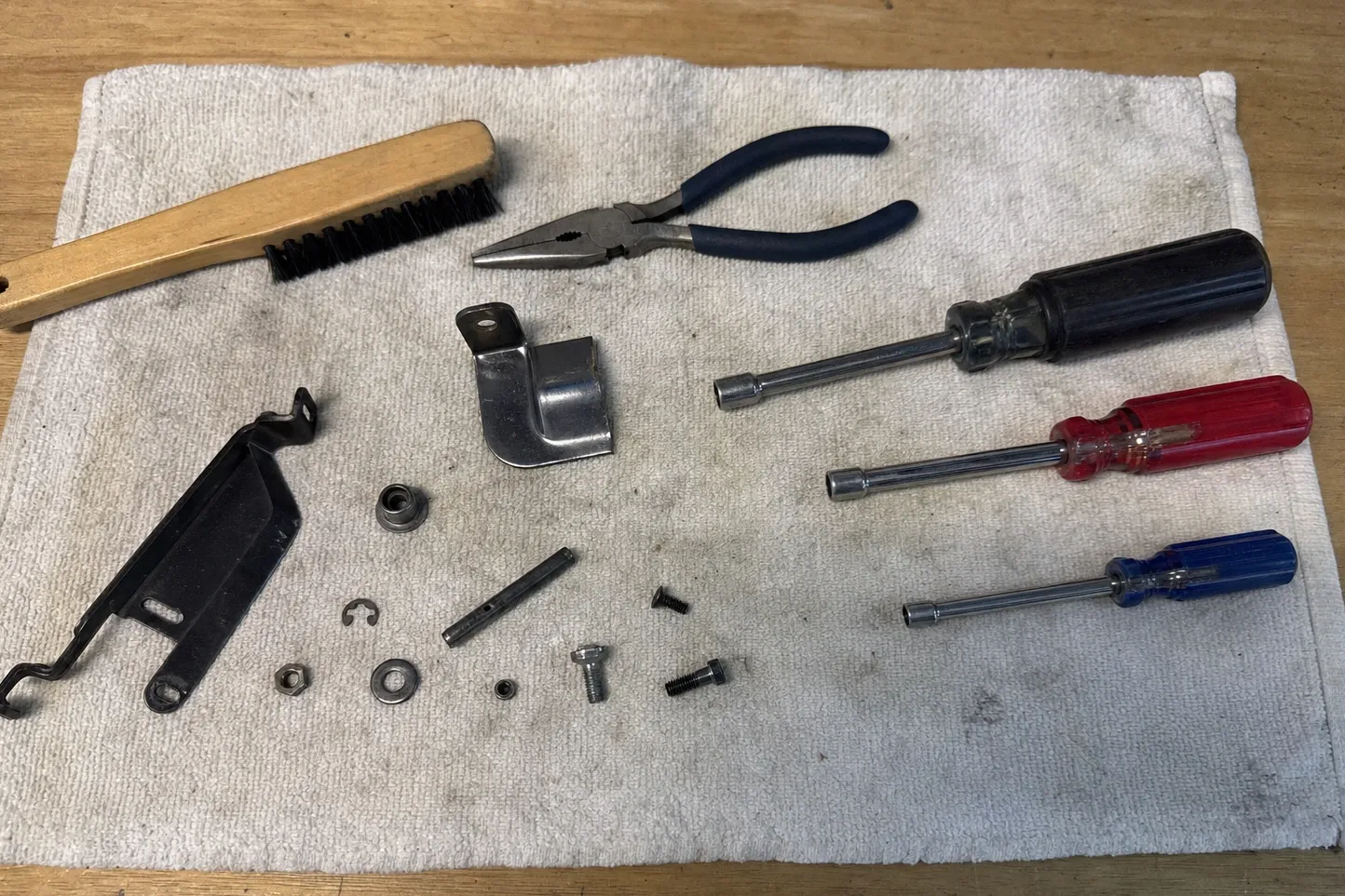 Two black formed brackets, one carrying a wire arm, with a bright steel cover, a bushing, an E-clip, a roll pin and loose screws beside blue-handled pliers and three nut drivers