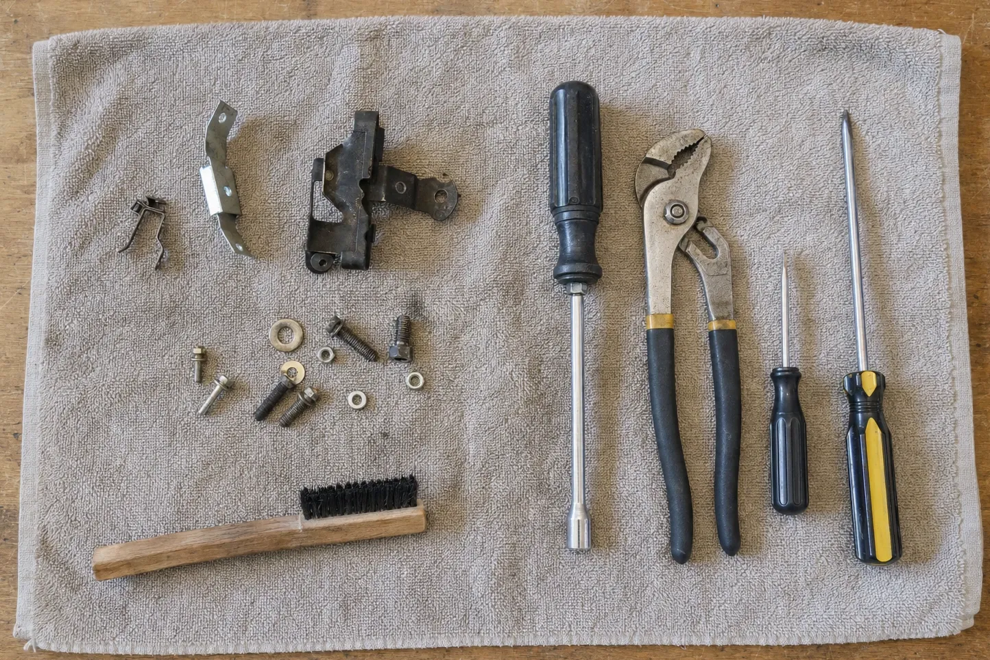 A blackened cam and latch body with a curved stainless bracket, a spring clip, washers and bolts, laid out with water-pump pliers, a nut driver, a pick and a large flat driver